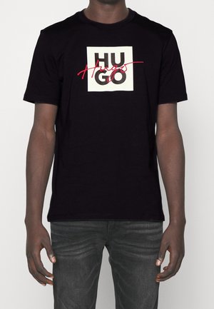 Man wearing a black Hugo Boss t-shirt with "HUGO" in bold black letters and "Hugo" in red script over a white square.