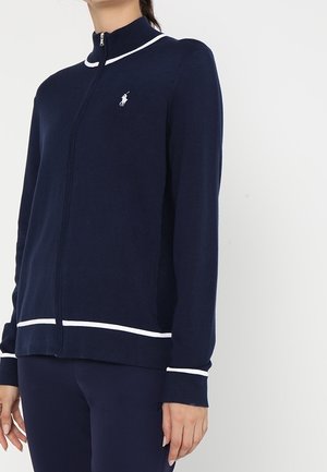 Femme portant une veste zippée bleu marine avec une bordure blanche et un petit logo brodé sur la poitrine, associée à un pantalon bleu marine assorti.