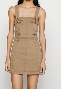 Tan denimklänning med fyrkantig halsringning, breda axelremmar och knäppta bröstfickor. Har en figurnära midja och minikorta nederdel.