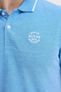 Lyseblå poloshirt med strukturvev, med en hvit brodert logo som sier "DENIM BLEND WITH ATTITUDE" på brystet.