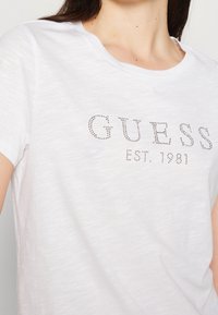Bílá bavlněná tričko s kulatým výstřihem, krátkými rukávy a třpytivým potiskem "GUESS EST. 1981" z rhinestones na přední straně.
