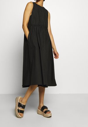 Person wearing a sleeveless black midi dress and black platform sandals standing against a plain light background.