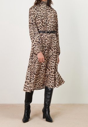 Kvinde iført en leopardprintet midi-kjole med lange ærmer, sort bælte og sorte knæhøje støvler med hæl, stående på et beige gulv.
