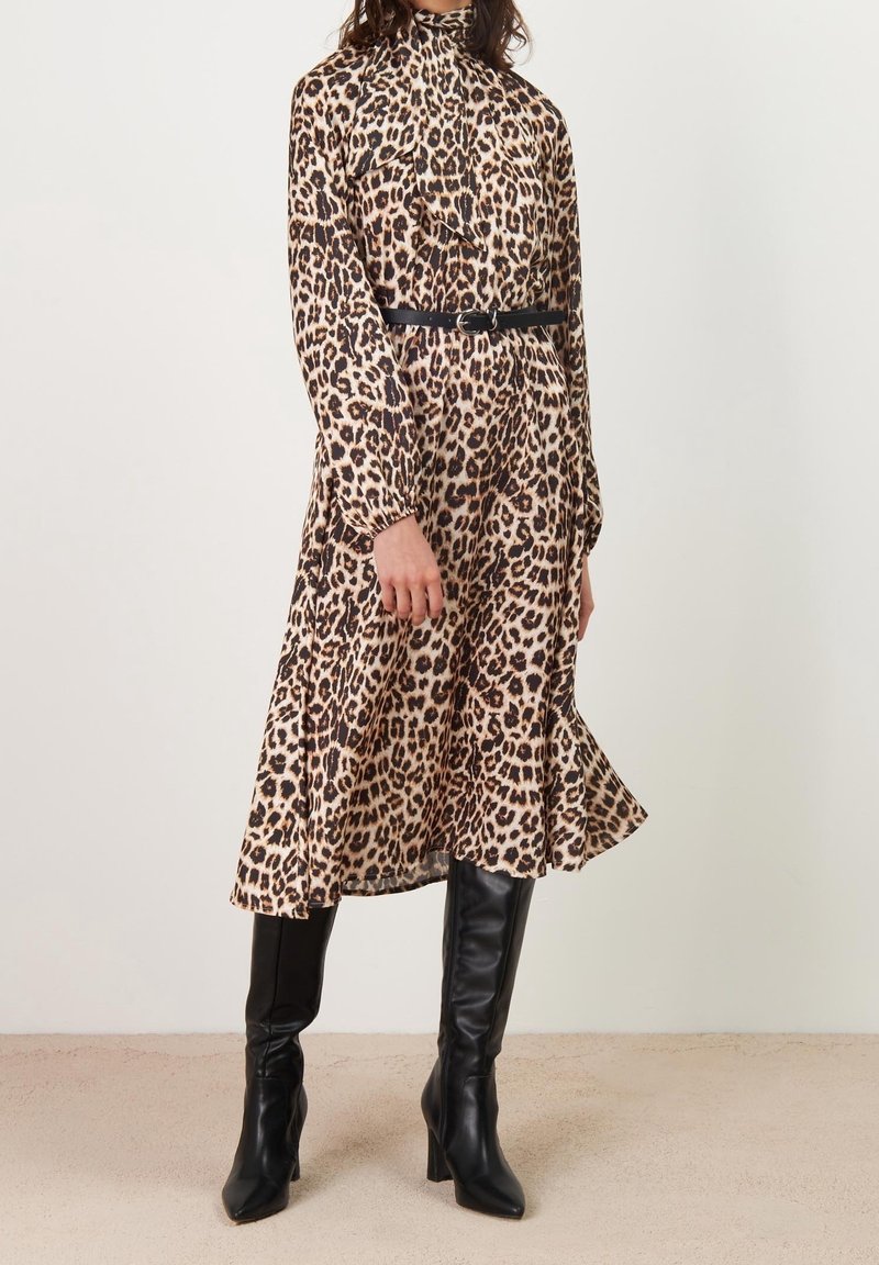 Femme portant une robe midi à manches longues à imprimé léopard, une ceinture noire et des bottes noires montantes à talons, debout sur un sol beige.