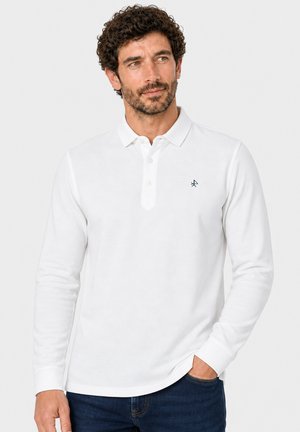 Hombre con cabello rizado que lleva una camisa polo blanca de manga larga y vaqueros azul oscuro, de pie con una mano en el bolsillo.
