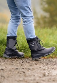 Zwarte waterdichte laarzen met een gevoerde kraag, veters en een gestructureerde rubberen zool, getoond op een persoon die over een grindpad wandelt.