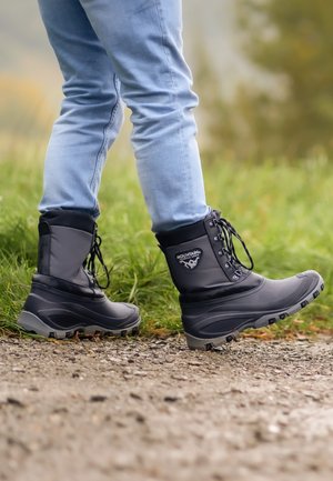 Schwarze wasserfeste Stiefel mit gepolstertem Kragen, Schnürsenkeln und strukturiertem Gummisohle, die an einer Person gezeigt werden, die auf einem Kiesweg läuft.