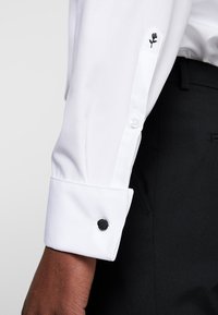 Camicia bianca a maniche lunghe con ricamo di una rosa nera sul polsino, caratterizzata da una texture liscia e dettagli di bottoni neri. Indossata con pantaloni neri.