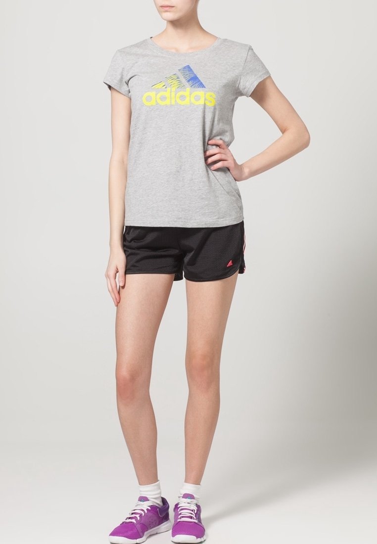 Grå kortärmad t-shirt med en blå och gul Adidas-logotyp, parat med svarta shorts. Modellen bär lila träningsskor och vita strumpor.
