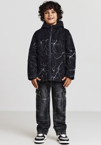 Giacca imbottita nera con motivo a saetta bianco, zip frontale e cappuccio; abbinata a pantaloni cargo scuri e scarpe da ginnastica bianche e nere.