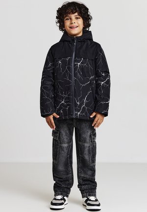 Giacca imbottita nera con motivo a saetta bianco, zip frontale e cappuccio; abbinata a pantaloni cargo scuri e scarpe da ginnastica bianche e nere.