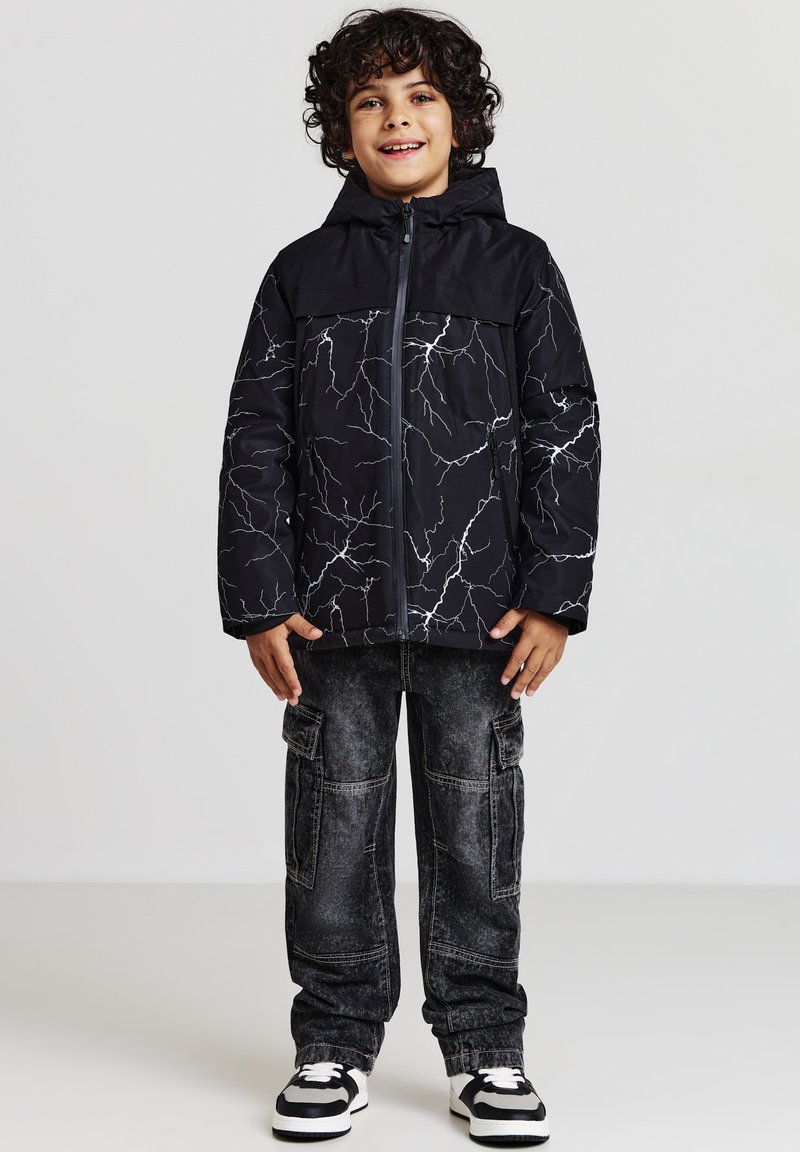 Giacca imbottita nera con motivo a saetta bianco, zip frontale e cappuccio; abbinata a pantaloni cargo scuri e scarpe da ginnastica bianche e nere.