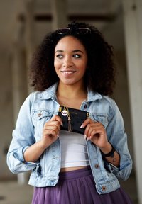 Černá peněženka s vyraženými vzory a zlatými kovovými doplňky, kterou drží osoba v džínové bundě a fialové sukni, se střapcem na klíče.