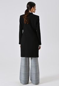 Cappotto lungo nero con silhouette strutturata, abbinato a pantaloni a gamba larga grigio a scacchi. Tessuto liscio con dettagli sartoriali.