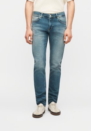 Άτομο που φορά στενά μπλε τζιν και μπεζ αθλητικά παπούτσια, στέκεται μπροστά σε λευκό φωτεινό φόντο.