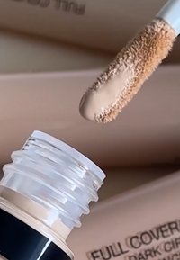 Flüssiger Concealer in einem transparenten Schlauch mit Applikator in Form eines Rehkitzfußes und pfirsichfarbener Formel vor einem beigen Hintergrund.