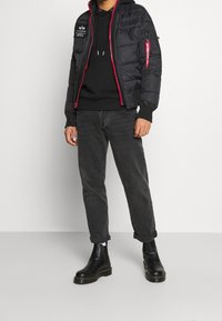 Giacca puffa nera con zip e dettagli rossi, dotata di cappuccio, tasca con zip angolata sulla manica, indossata sopra un hoodie nero e jeans scuri.