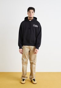 Haut à capuche noir avec une poche avant et un design graphique blanc, associé à un pantalon beige et des baskets jaunes avec des accents noirs sur un sol jaune.