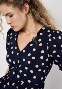 Navyblaues Kleid mit großen cremefarbenen Punkten, kurzen Puffärmeln, V-Ausschnitt und Knopfleiste; leichter Stoff mit fließender Silhouette.