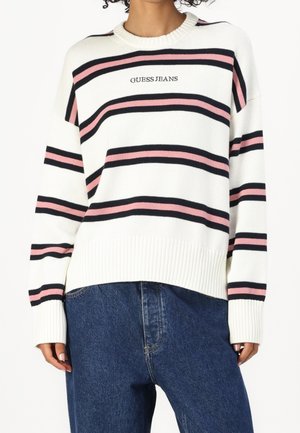 Biały sweter dziany z różowymi i czarnymi poziomymi paskami oraz napisem „GUESS JEANS”, noszony z niebieskimi dżinsami.