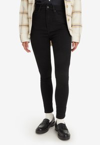 Personne portant un jean skinny noir taille haute, des chaussettes blanches, des mocassins noirs et une veste en tartan crème avec des rayures beige et marine.