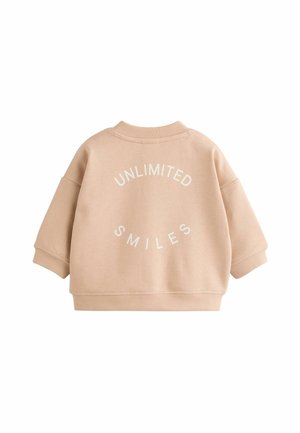 Vaaleanbeige huppari, jossa on ribattua kaula-aukkoa ja laskettuja olkapäitä. Takana on valkoisella teksti "UNLIMITED SMILES".
