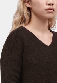 Femme aux longs cheveux blonds portant un pull en maille à col en V marron foncé sur un fond clair uni.