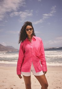 Pink linen button-up shirt with a chest pocket, relaxed fit, paired with white shorts; model stands on a beach with ocean background.