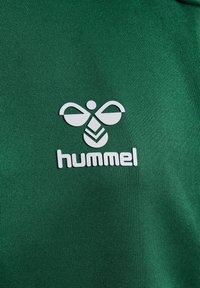 Camiseta atlética verde con una textura suave que presenta un logo blanco que se asemeja a una abeja y la palabra "hummel" en letras negritas.
