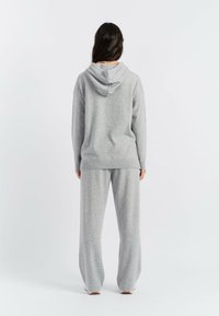 Grauer Strickpullover mit Kapuze, lockerer Schnitt und gerippten Bündchen, kombiniert mit passenden grauen weiter geschnittenen Hosen, von hinten gesehen.