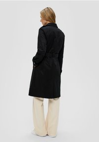 Manteau noir avec une ceinture, doté d'un col et de manches longues. Porté par-dessus un pantalon large de couleur crème.