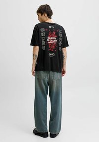 Zwart graphic T-shirt met gedurfde rode en witte tekst en illustraties op de achterkant. Gecombineerd met losse, vervaagde blauwe denim broeken en zwarte schoenen.