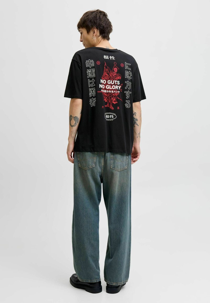 Sort grafisk t-shirt med fed rød og hvid tekst og illustrationer på ryggen. Sammen med løse, falmede blå denimbukser og sorte sko.