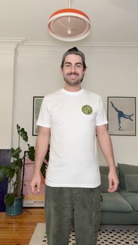 Vit bomullst-shirt med en grön och gul rund logotyp på vänstra bröstet, kombinerad med gröna manchesterbyxor. Inomhusmiljö.