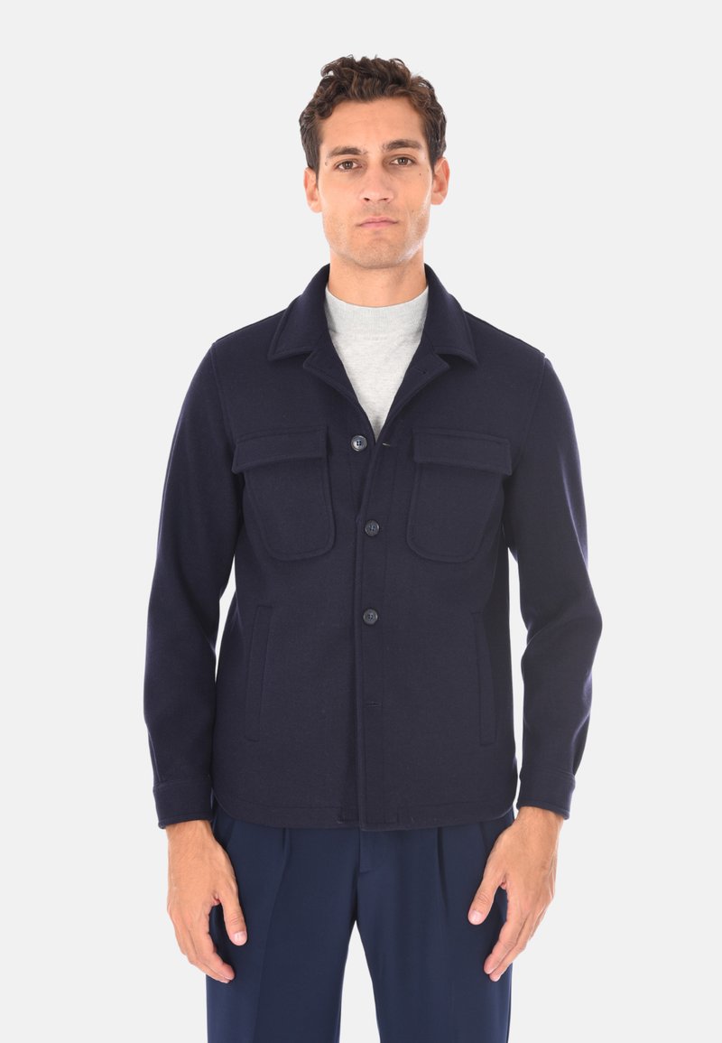 Giacca in misto lana blu navy con collo, quattro tasche, chiusura con bottoni e texture morbida. Indossata sopra una camicia grigia chiaro.