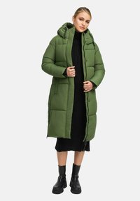 Kvinna i en lång grön vadderad kappan med huva över en svart ribbad klänning och svarta kraftiga ankelstövlar, stående mot en vit bakgrund.