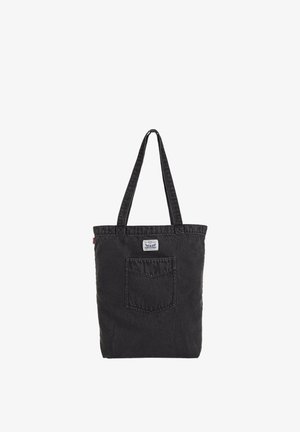 Sort denim tote taske med lange håndtag, frontlomme og lille hvid label over lommen på en ensfarvet baggrund.