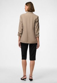 Blazer beige su misura con maniche a tre quarti, indossato sopra pantaloni neri fino al ginocchio. Tessuto liscio, rever classico e design a taglio dritto.