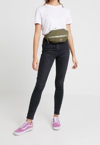 Femme portant un t-shirt blanc, un jean skinny noir, des baskets roses et une banane vert olive avec une fermeture éclair, se tenant devant un fond uni.