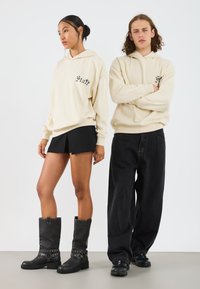 Felpe beige con la scritta "Staff", pantaloni corti neri e stivali neri al ginocchio con dettagli a fibbia. Jeans scuri con vestibilità rilassata.