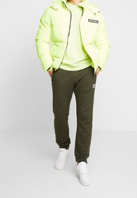 Neongrön puffjacka med frontdragkedja och huva, kombinerad med olivgröna sweatpants med en vit logotyp och vita sneakers.