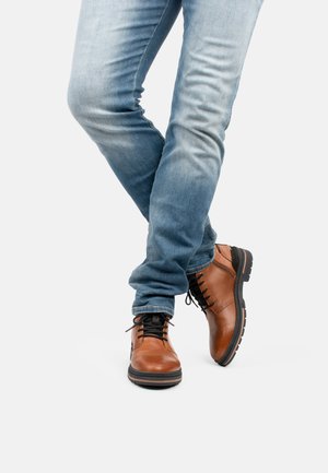 Brown leather ankle boots with black laces and rubber soles, worn with blue jeans. Boots feature a smooth finish and contrasting sole.