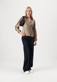 Blusa envolvente de seda em bege com mangas bufantes, combinada com calças de ganga escuras de perna larga. Modelo a usar uma elegante mala preta de ombro e sapatos brancos.
