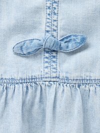 Lyseblå denimstoff har en knyttet sløyfe og rynket volangdetalj langs hemmet, med synlige sømmer.