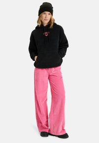 Felpa nera in pile con logo ricamato rosso, abbinata a pantaloni in velluto a costo rosa a gamba larga e scarpe nere senza lacci. Accessorio berretto nero.