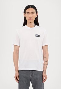 Baltas kokvilnas T-krekls ar apaļu kaklu un īsām piedurknēm. Uz kreisās krūts daļas ir neliels melns taisnstūra logotips.