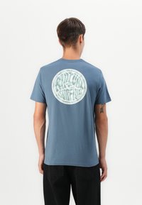 Blå bomulls t-skjorte med et rundt grafisk design i hvitt på baksiden, satt mot en enkel bakgrunn.