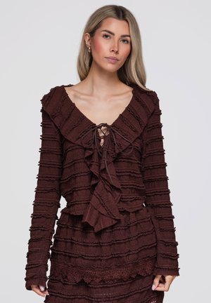 Jeune femme aux longs cheveux blonds portant un haut texturé à manches longues marron foncé avec des volants et un lien au niveau du col.