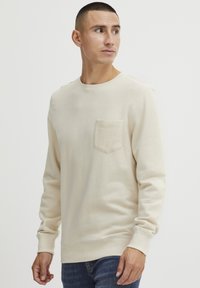 Sweatshirt beige en tissu doux avec des manches longues, un col rond et une poche avant. Présente des poignets et un ourlet côtelés pour un look ajusté.