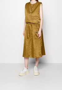 Olive green satin dress with a sleeveless design, front zip closure, and a wide, flowing skirt. Paired with white platform sneakers.